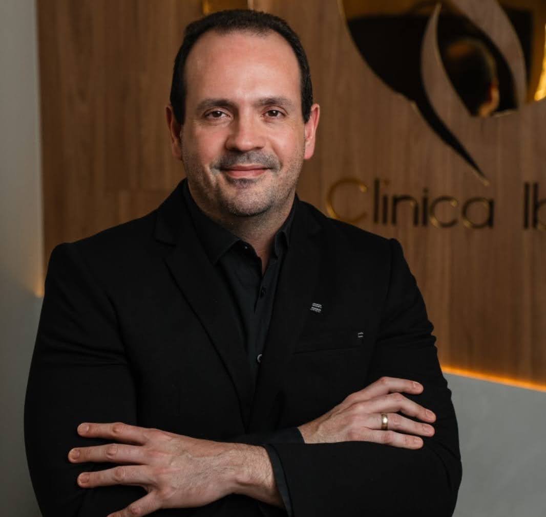 Homem de blazer preto, de braços cruzados, diante da parede com o nome da clínica
