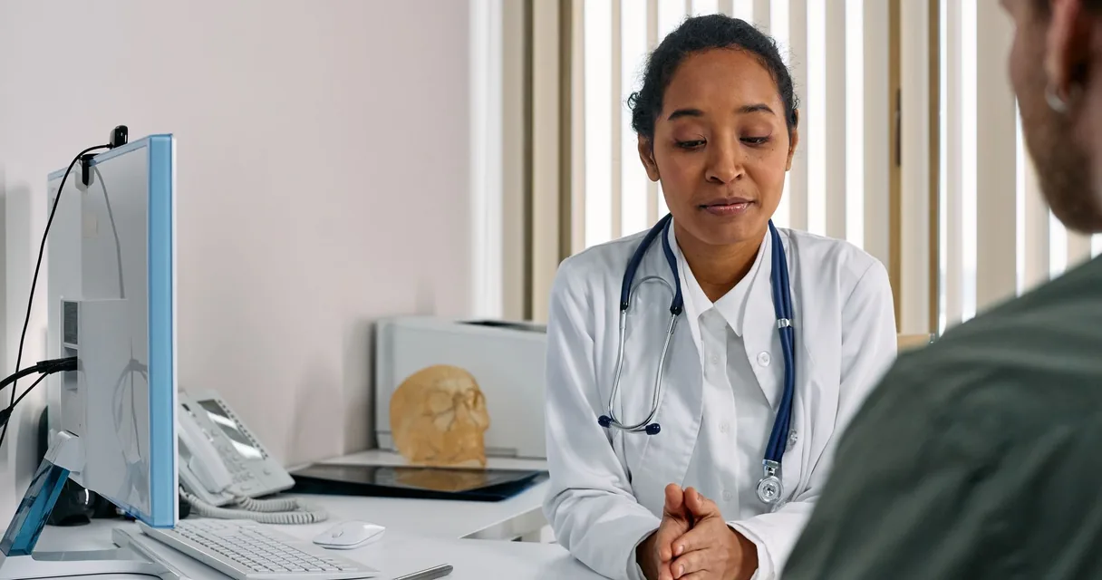 Médica de jaleco branco atendendo paciente em consultório, com computador e crânio de estudo sobre a mesa